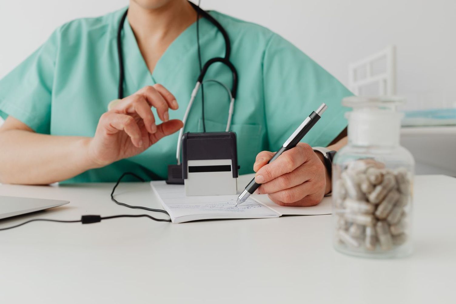 Healthcare professional writing a prescription beside a stethoscope and a jar of pills.