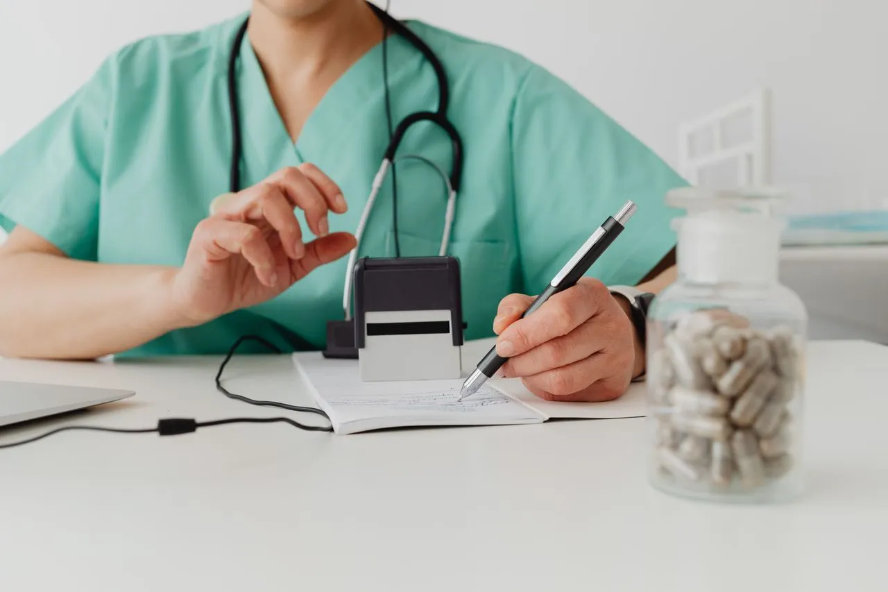 Healthcare professional writing a prescription beside a stethoscope and a jar of pills.