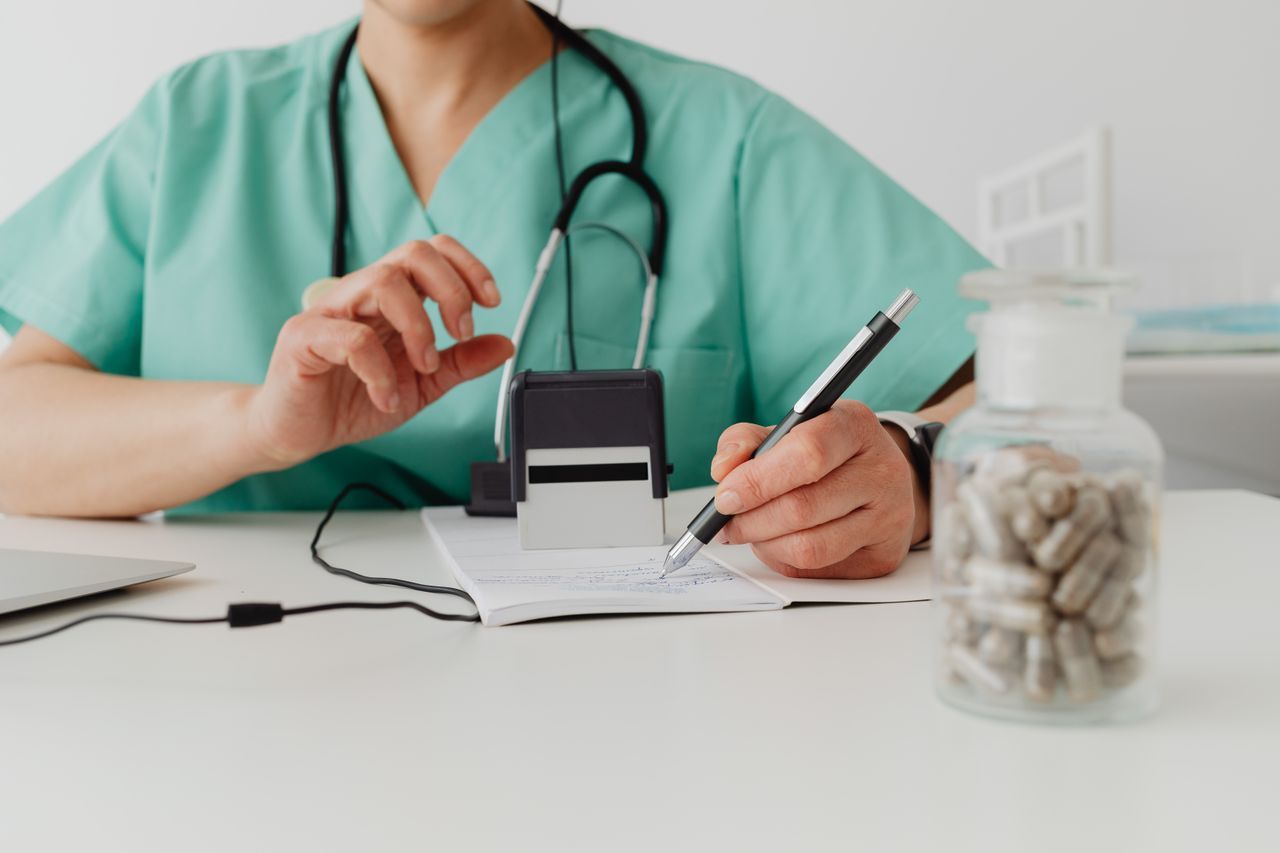 Healthcare professional writing a prescription beside a stethoscope and a jar of pills.
