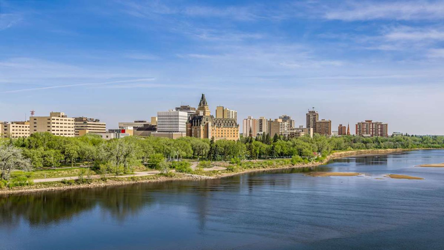 Skyline of Saskatoon, Saskatchewan, along the South Saskatchewan River under a clear blue sky.