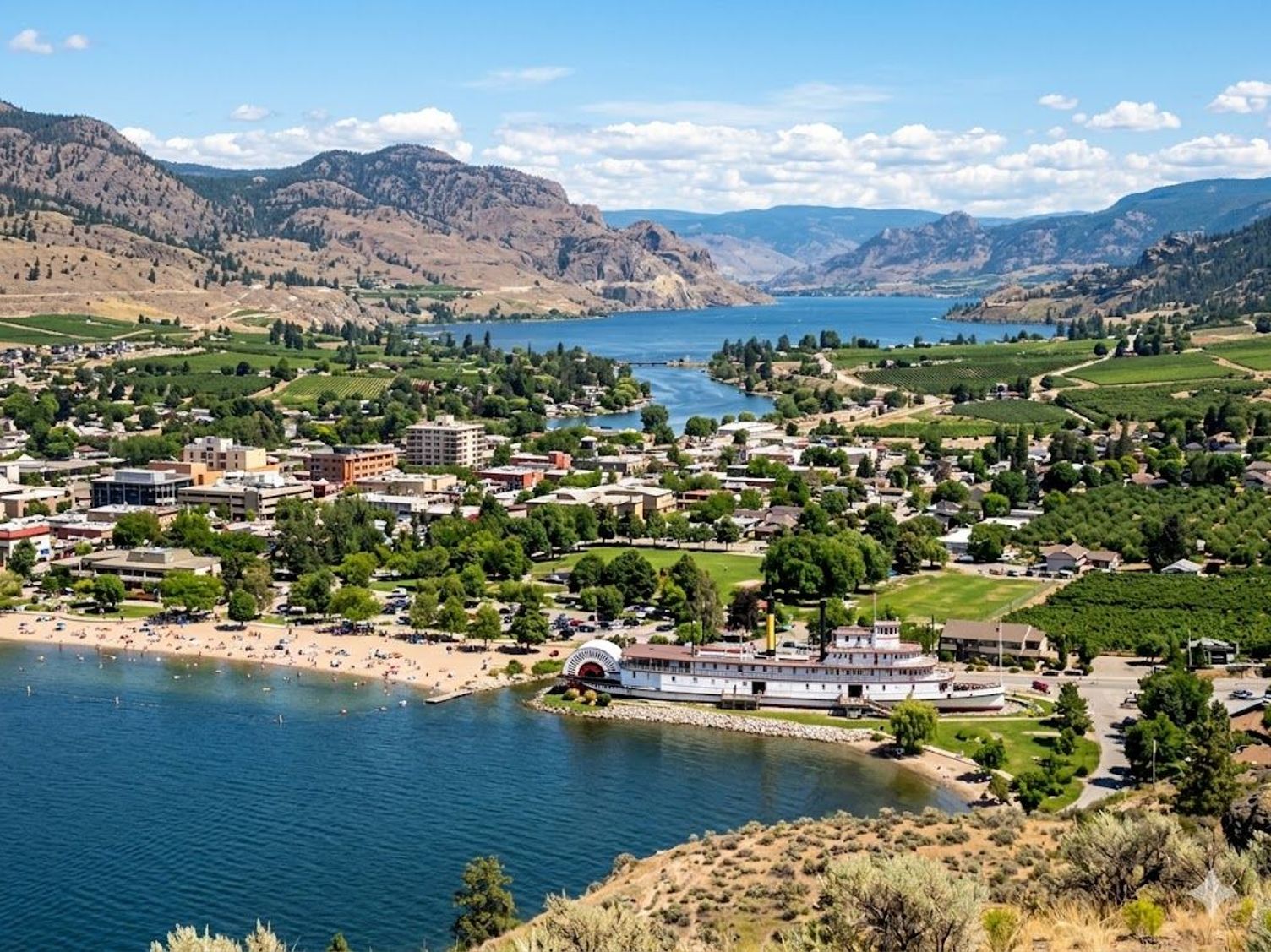 Aerial view of Penticton, BC, showing Okanagan Lake, a sandy beach, and the historic SS Sicamous paddlewheeler.