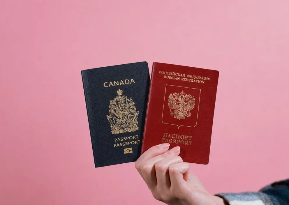 A hand holding a Canadian passport and a Russian passport side-by-side against a pink background.