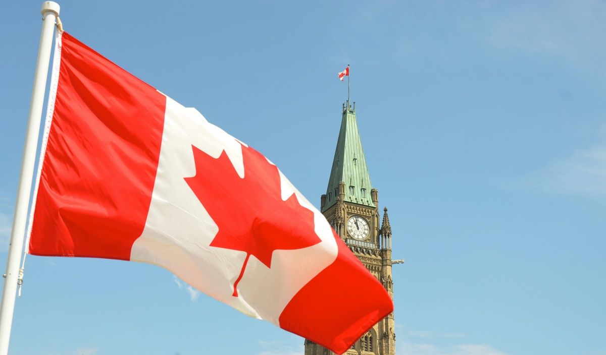 A Canadian flag waving in front of the Peace Tower in Ottawa under a clear blue sky.