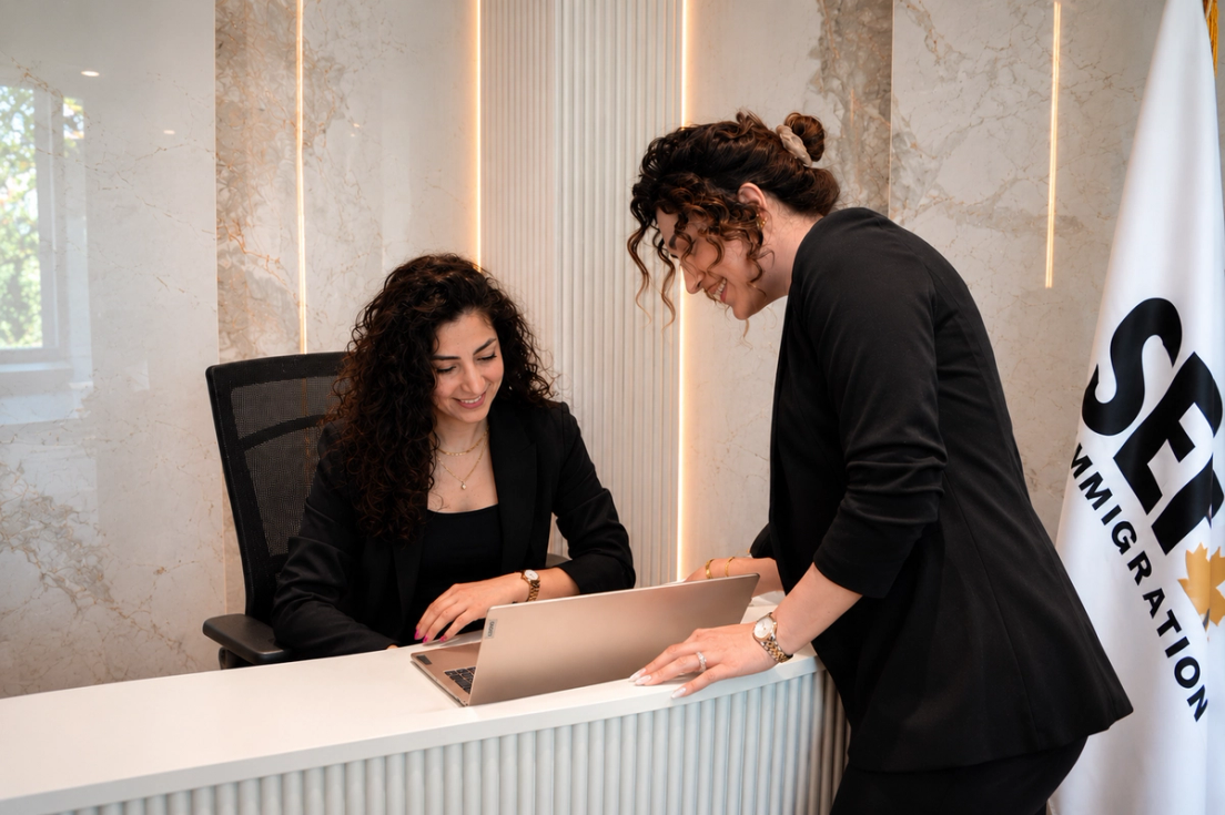 Two women in black blazers looking at a laptop computer together at an office desk.