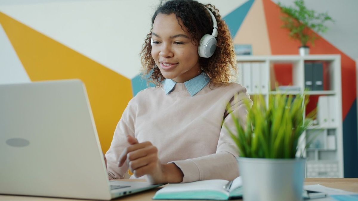 A young woman wearing headphones works on a laptop at a desk with an open notebook.