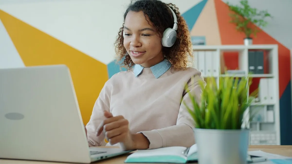 A young woman wearing headphones works on a laptop at a desk with an open notebook.