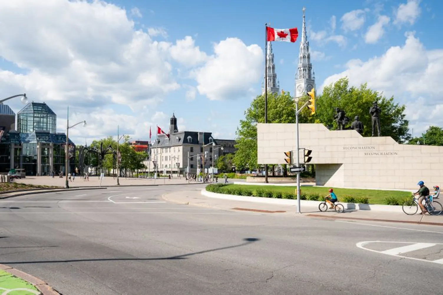Street scene in Ottawa featuring the Peacekeeping Monument, a Canadian flag, and cyclists on a sunny day.