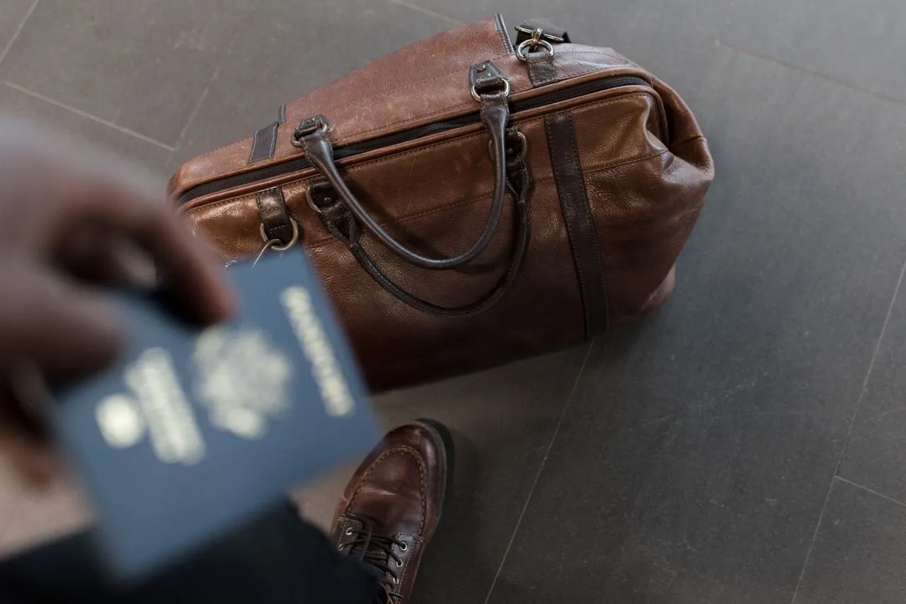 A traveler holds a passport beside a brown leather bag and wears brown shoes on a tiled floor.