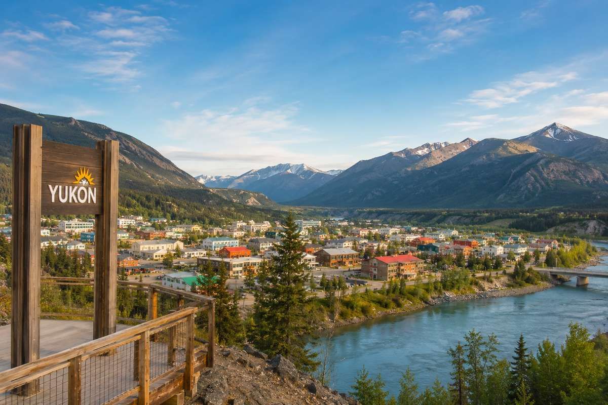 A wooden Yukon sign stands on an overlook with a town along a river surrounded by mountains.