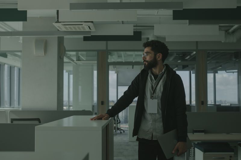 Bearded man wearing an ID badge stands in a modern office, holding a folder beside a counter.