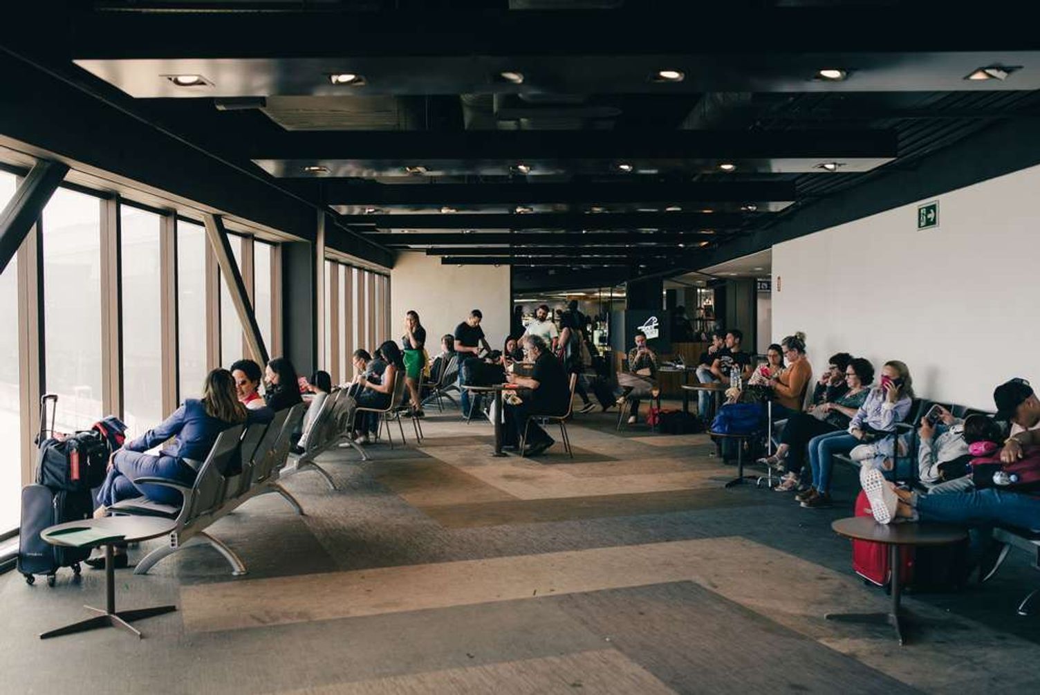 Travelers waiting with suitcases in a spacious airport terminal lounge next to large windows.