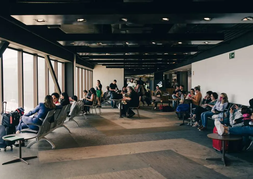 Travelers waiting with suitcases in a spacious airport terminal lounge next to large windows.