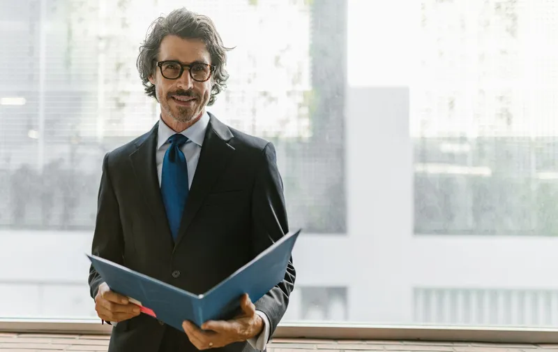 A man wearing a dark suit, tie, and glasses stands near a window holding an open blue folder.