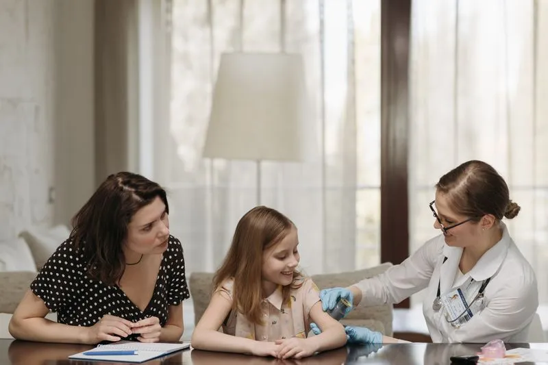 A doctor in a white coat examines a young girl's arm while her mother watches.