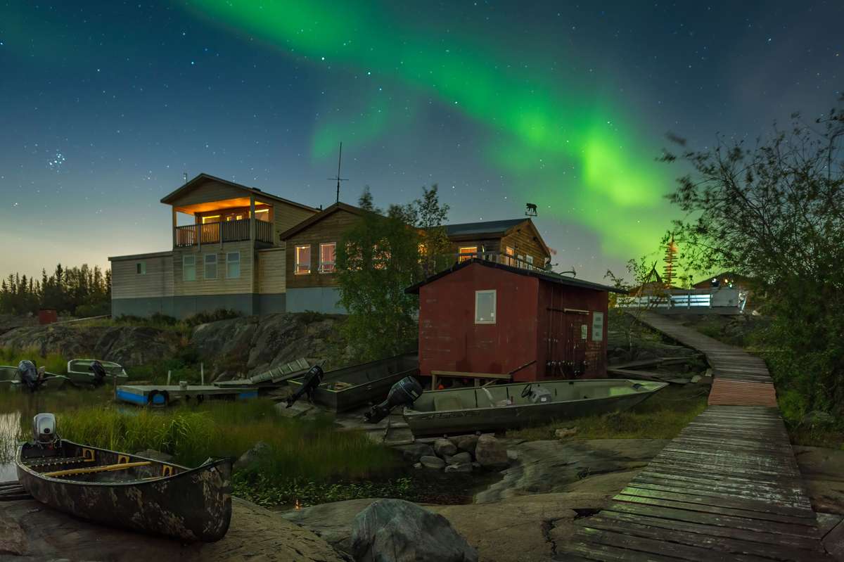 Green northern lights glowing above wooden cabins and small boats along a shore at night.