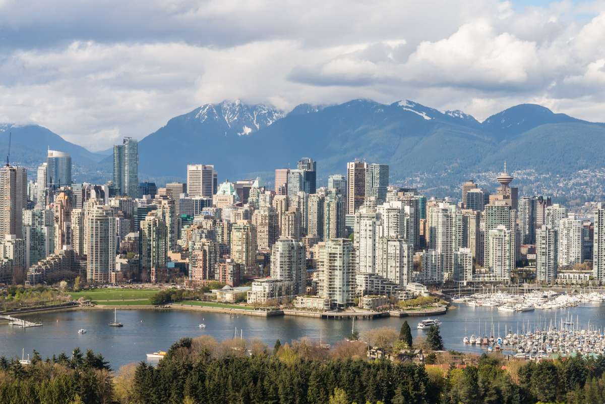 Skyline of Vancouver, British Columbia, featuring skyscrapers, a waterfront harbor, and snow-capped mountains.