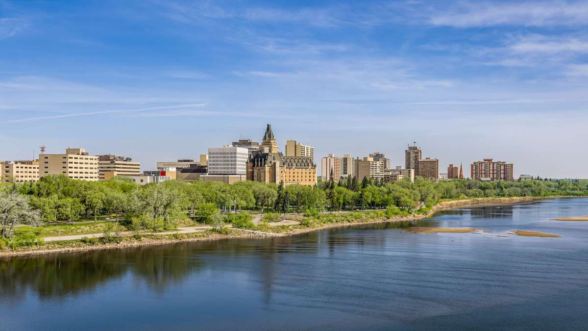 Skyline of Saskatoon, Saskatchewan, along the South Saskatchewan River under a clear blue sky.