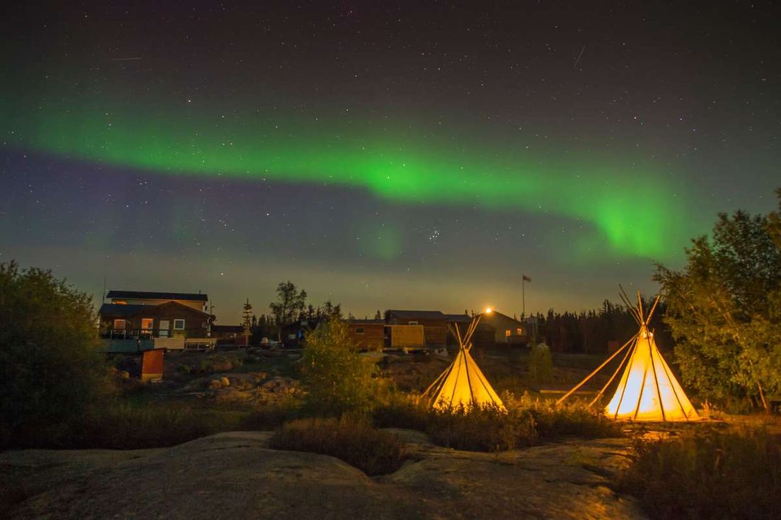 Green aurora borealis glows in the night sky above illuminated tipis and small wooden buildings.