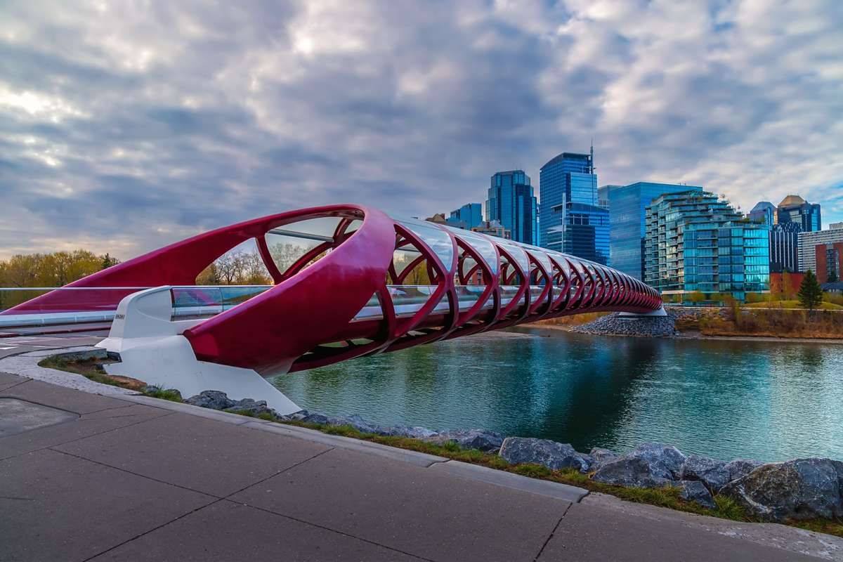 The red Peace Bridge spans the Bow River leading to downtown Calgary skyscrapers under a cloudy sky.