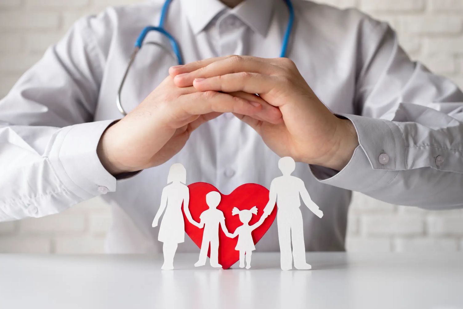 Close-up of a doctor with a stethoscope, hands forming a protective gesture over paper cutouts of a family in front of a red heart, symbolizing healthcare and social services protection.