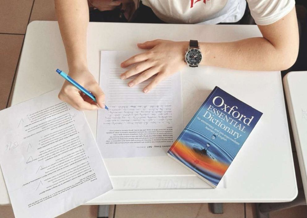 Overhead view of a person writing an exam at a desk next to an Oxford dictionary.