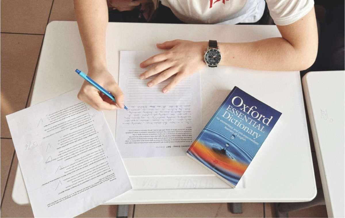 Overhead view of a person writing an exam at a desk next to an Oxford dictionary.