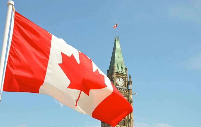 A Canadian flag waving in front of the Peace Tower in Ottawa under a clear blue sky.