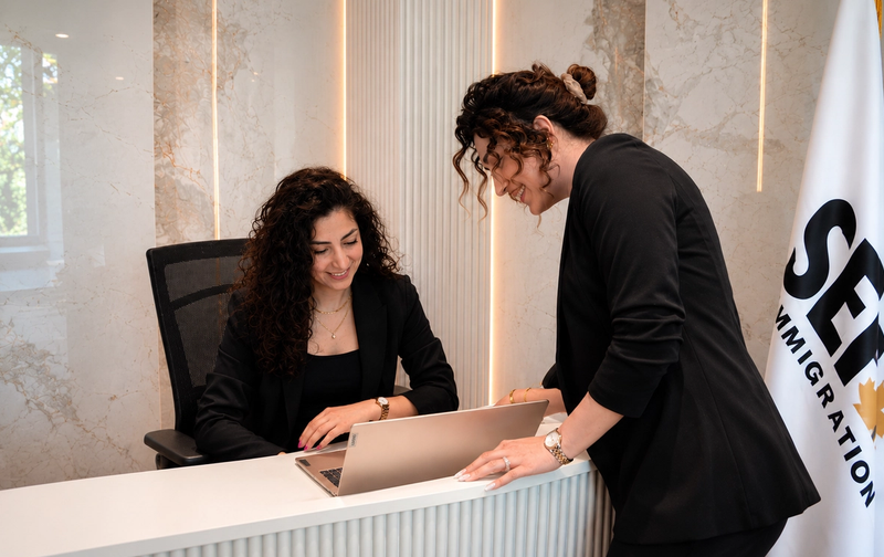 Two women in black blazers look at a laptop in an office with an SEP Immigration flag.