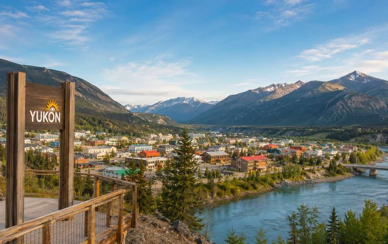 A wooden Yukon sign stands on an overlook with a town along a river surrounded by mountains.