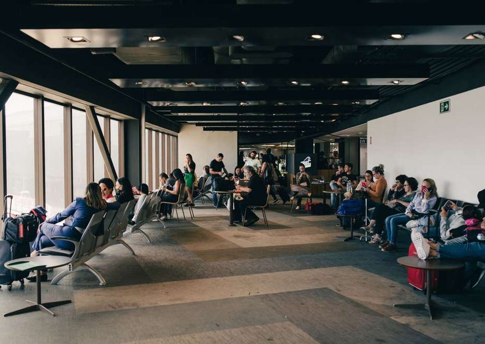 Travelers waiting with suitcases in a spacious airport terminal lounge next to large windows.