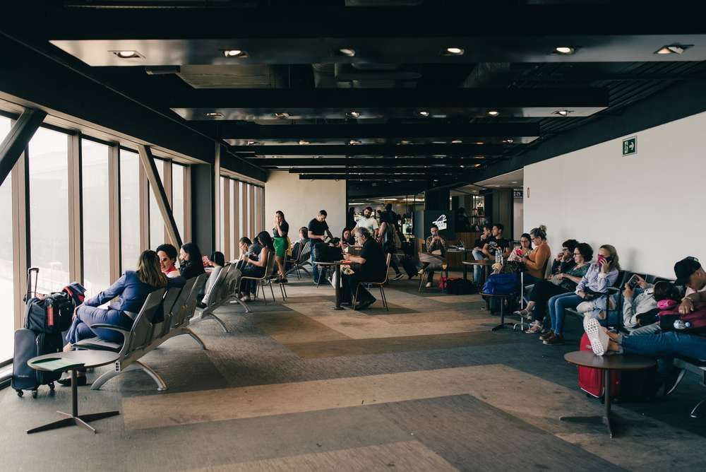 Travelers waiting with suitcases in a spacious airport terminal lounge next to large windows.