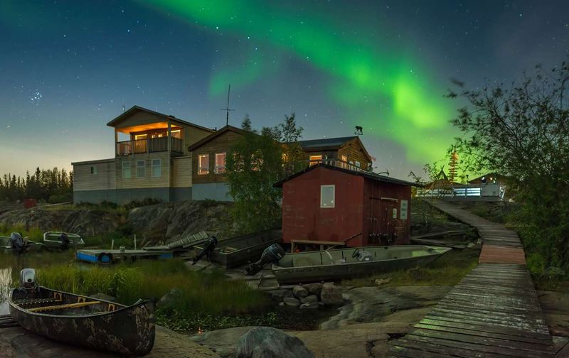 Green northern lights glowing above wooden cabins and small boats along a shore at night.