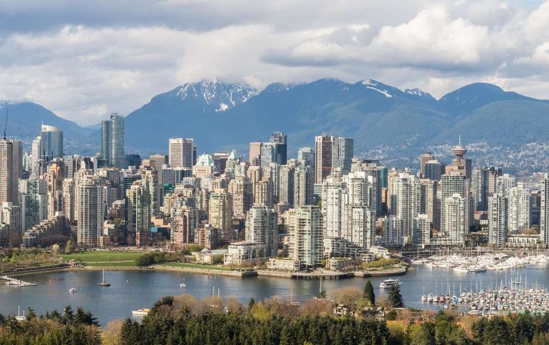 Skyline of Vancouver, British Columbia, featuring skyscrapers, a waterfront harbor, and snow-capped mountains.