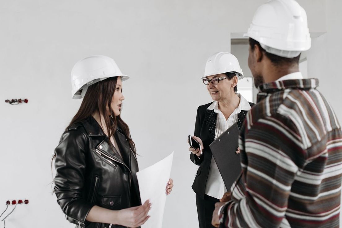 Three professionals wearing white hard hats discuss plans inside an unfinished building.