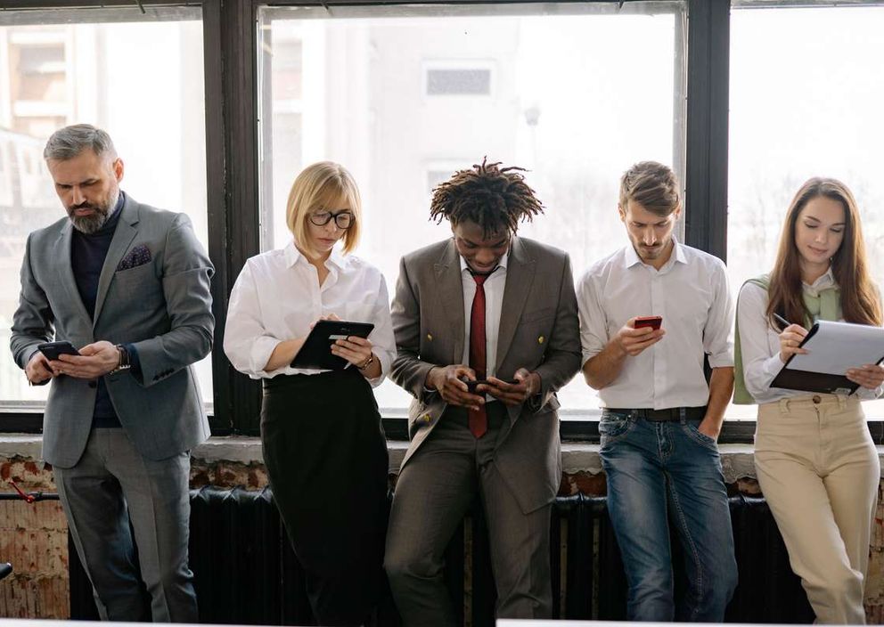 Five professionals standing in a row by a large window, using phones, a tablet, and a clipboard.