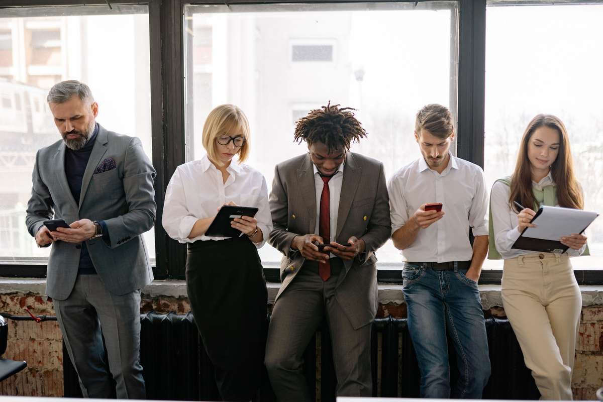 Five professionals standing in a row by a large window, using phones, a tablet, and a clipboard.