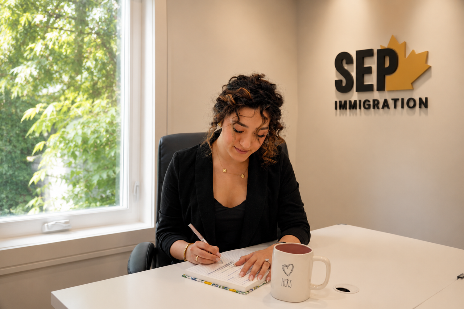 A woman writes in a notebook at an office desk with a SEP Immigration logo on the wall behind her.