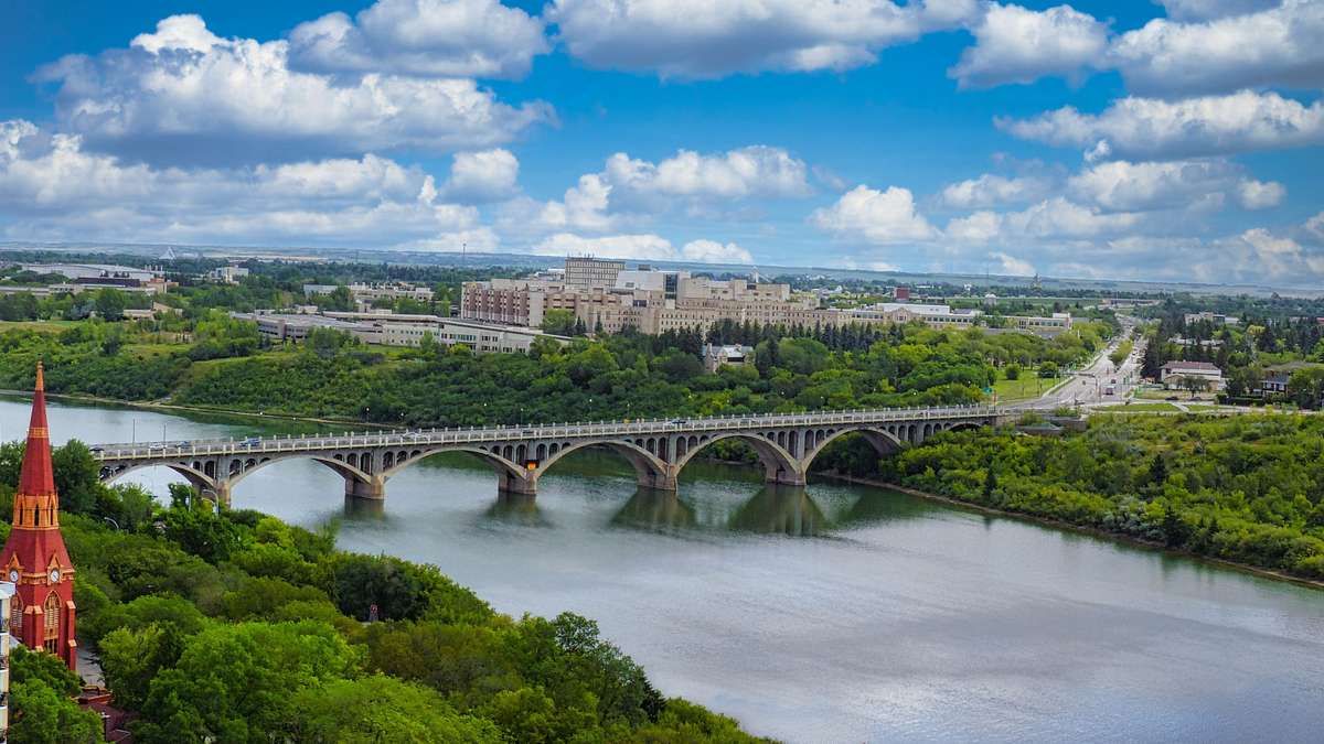 Aerial view of University Bridge crossing a wide river surrounded by green trees and buildings in Saskatoon.