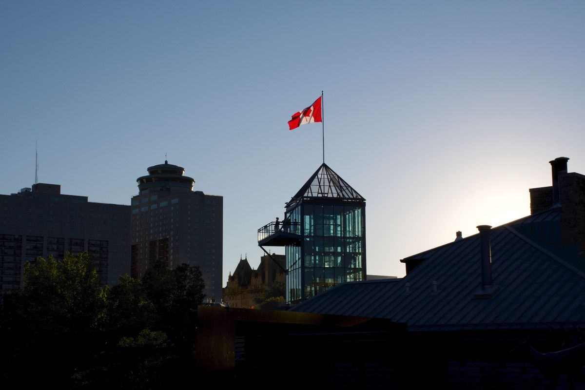A Canadian flag flying atop a modern glass tower against a clear sky in a city skyline.