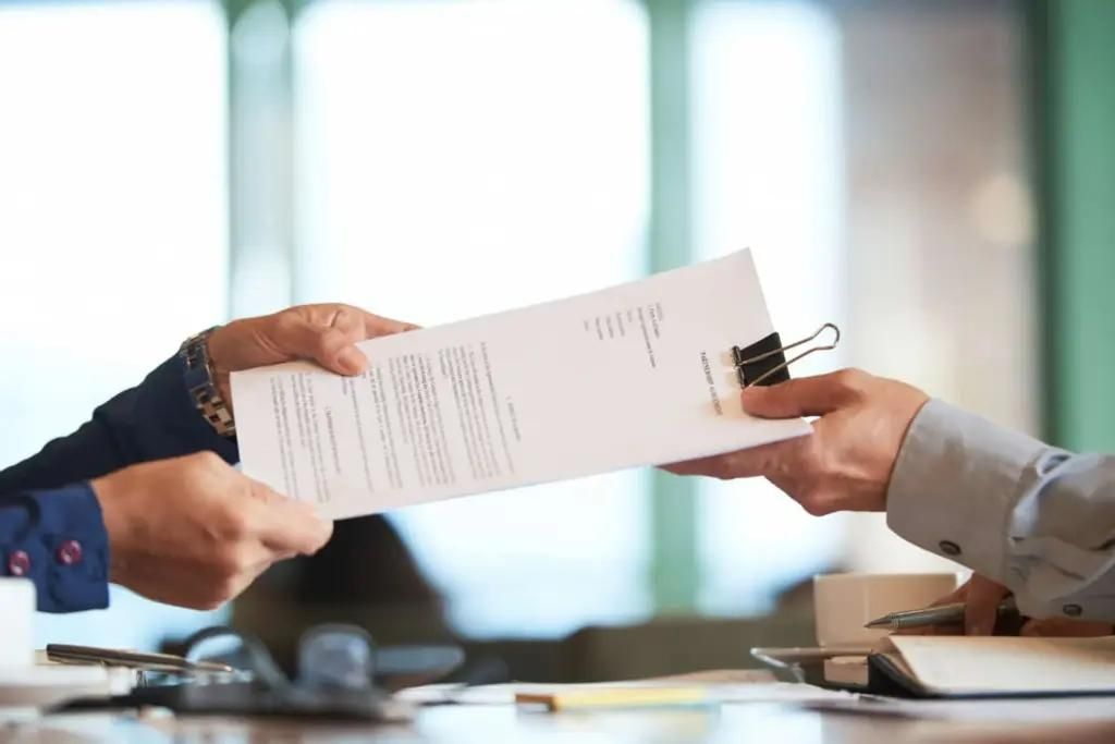 One person handing a stack of official documents clipped together to another person across a desk in a professional setting.