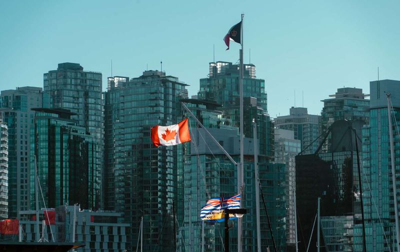 Canadian and British Columbia flags flying in front of modern glass high-rises in Vancouver.