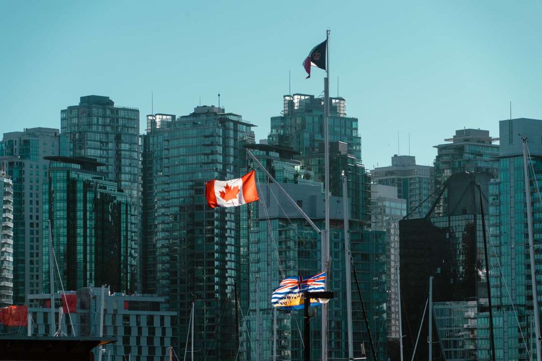 Canadian and British Columbia flags fly in front of modern high-rise buildings in Vancouver.