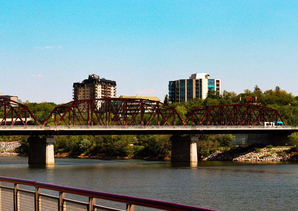 A red steel truss bridge crossing a river with city buildings and trees in the background under a blue sky.