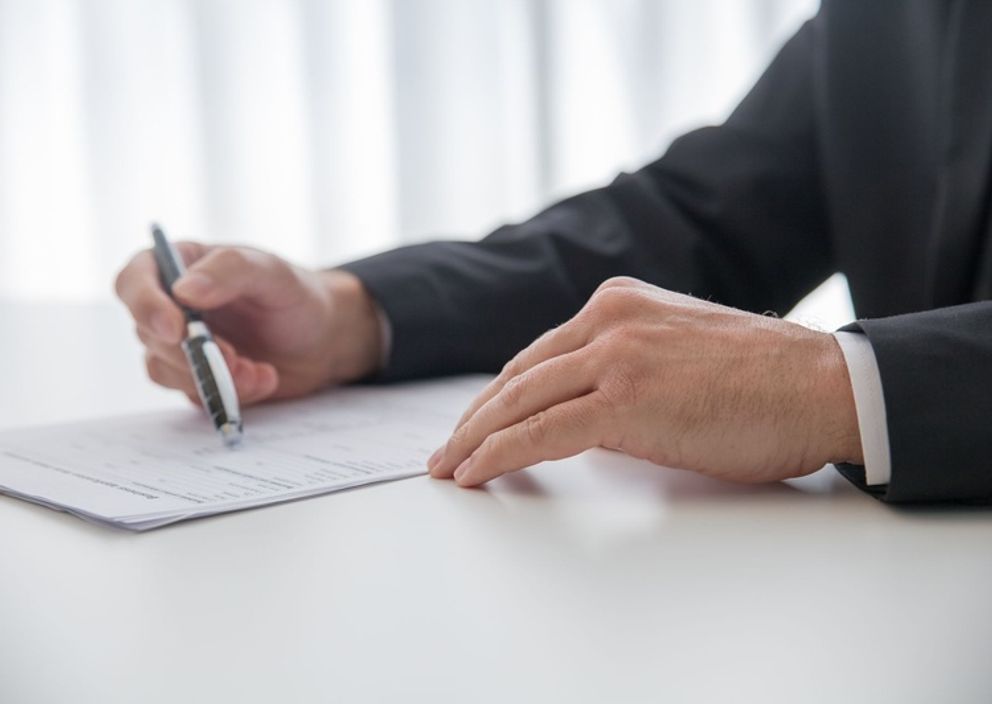 Close-up of a person in a suit holding a pen over a document on a white table.