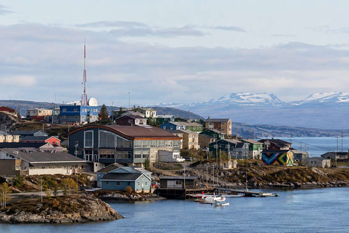 A northern town with colorful buildings, floatplanes at a dock, and snow-capped mountains across the water.