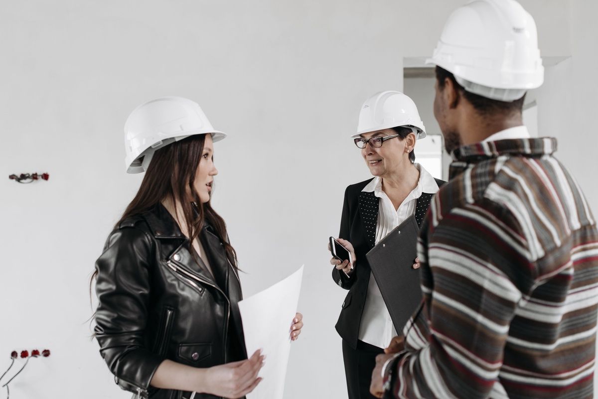 Three professionals wearing white hard hats discuss plans inside an unfinished building.