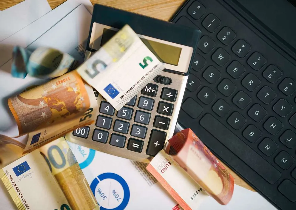 Euro banknotes, a calculator, financial charts, and a laptop keyboard on a desk.