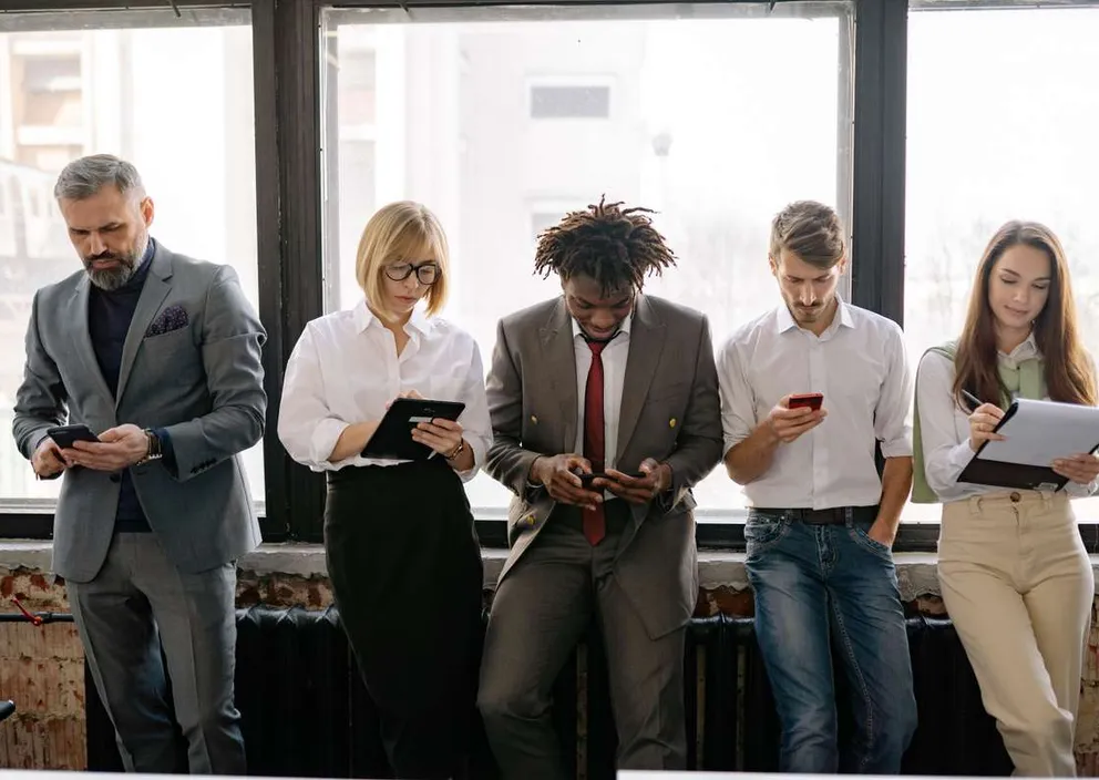 Five professionals standing in a row by a large window, using phones, a tablet, and a clipboard.