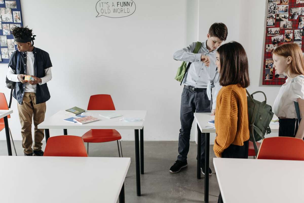 Students with backpacks standing around desks in a bright classroom.