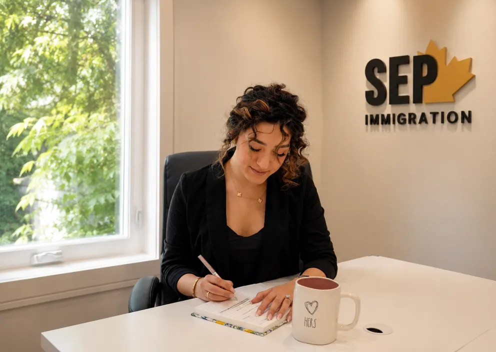 A woman writes in a notebook at an office desk with a SEP Immigration logo on the wall behind her.